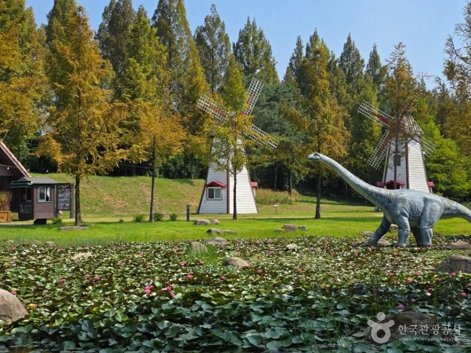 A long-necked dinosaur sculpture stands beside a water-lily pond with two white windmill structures and trees behind